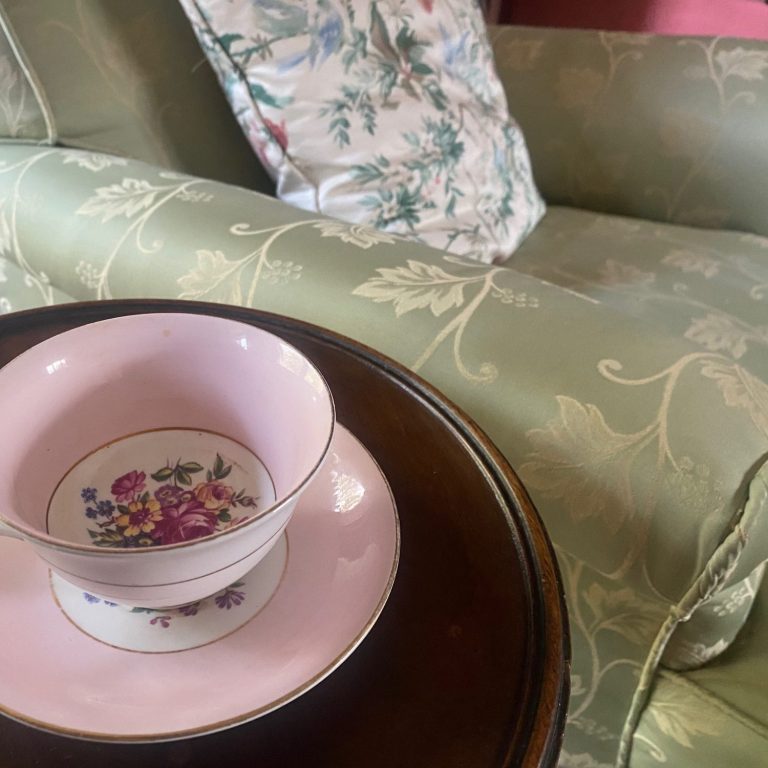 Pretty antique tea cup and saucer on a small table beside an elegant green satin covered antique armchair.