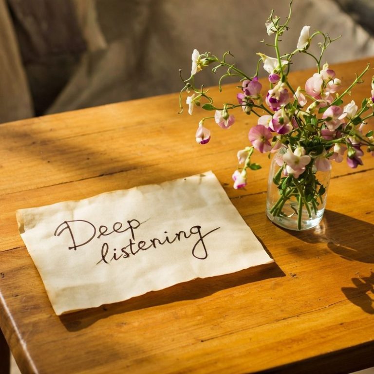 Honey coloured wooden table, small jar of garden of flowers, piece of parchment that says 'Deep Listening' in pen and ink.